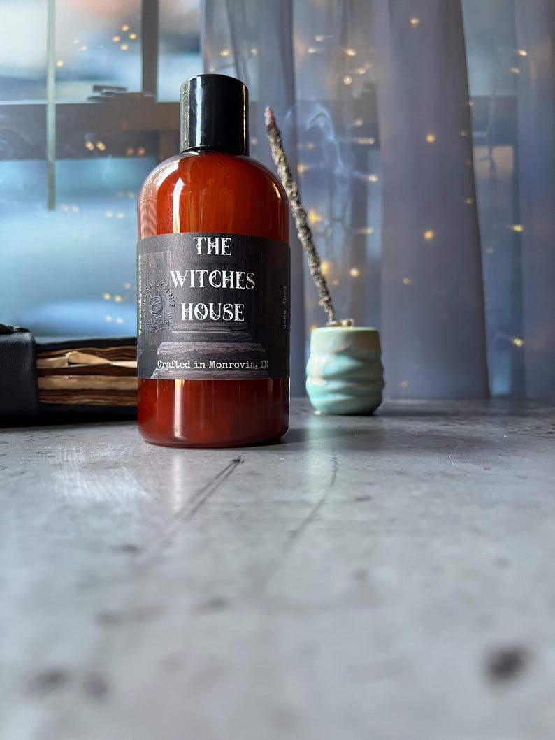 Puede incluir: Una botella cil&iacute;ndrica de pl&aacute;stico &aacute;mbar con tapa negra, que presenta una etiqueta negra con texto blanco que dice THE WITCHES HOUSE y Crafted in Monrovia, IN. La botella contiene un l&iacute;quido oscuro y est&aacute; colocada sobre una superficie gris.