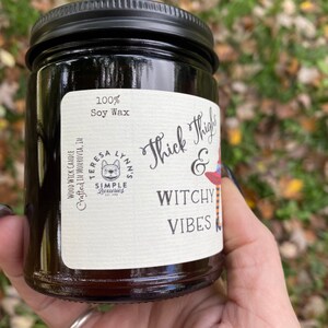 A hand holds a dark candle jar.  The label reads Thick Thighs & Witchy Vibes, wood wick soy candle.  The graphic is anime inspired witch legs with black and orange striped stockings, topped with red witch hat.  The background is a fall scene.