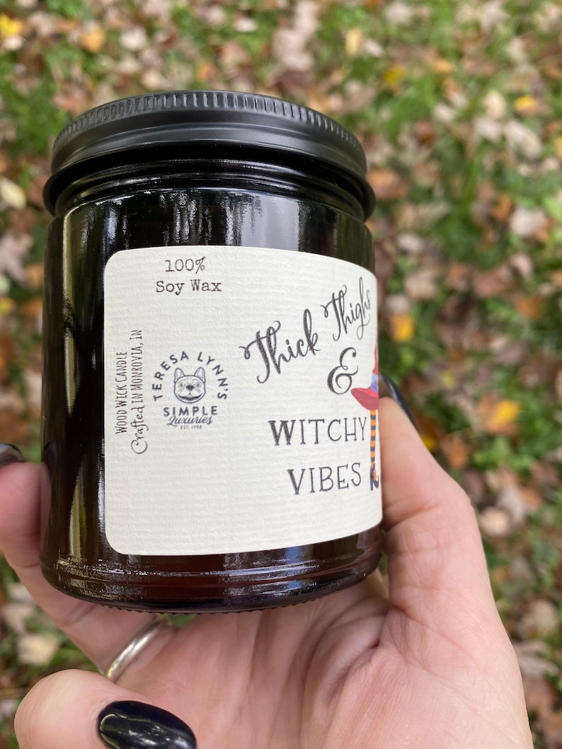 A hand holds a dark candle jar.  The label reads Thick Thighs & Witchy Vibes, wood wick soy candle.  The graphic is anime inspired witch legs with black and orange striped stockings, topped with red witch hat.  The background is a fall scene.