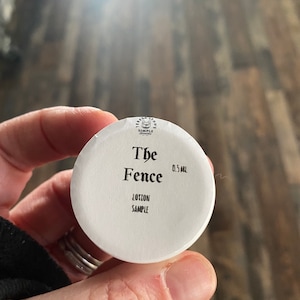 Op de afbeelding: Een kleine witte ronde deksel van een potje met zwarte tekst: Teresa Lynn's Simple Scents, The Fence, 0,5 ml en Lotion Sample. De deksel heeft een gladde, matte afwerking en een kleine afbeelding van een kattenkop in het midden bovenaan.