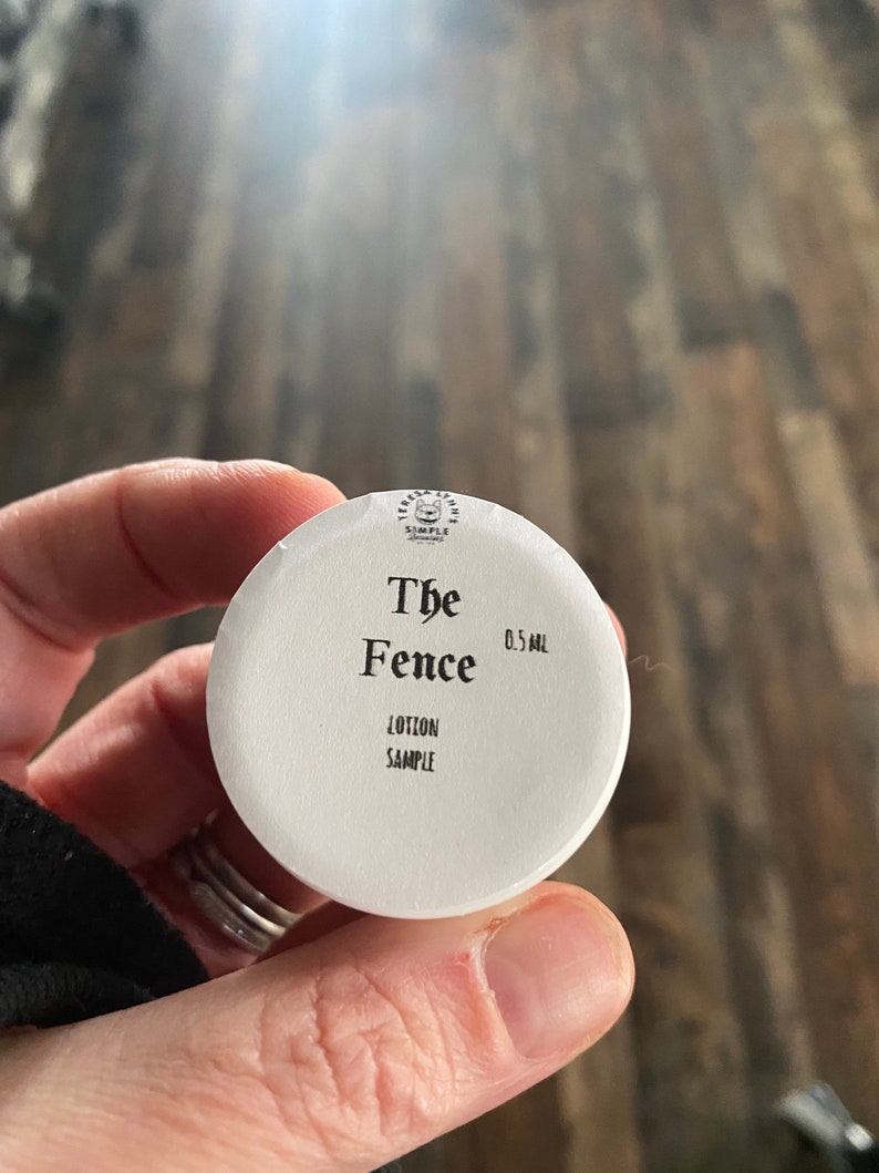 Op de afbeelding: Een kleine witte ronde deksel van een potje met zwarte tekst: Teresa Lynn's Simple Scents, The Fence, 0,5 ml en Lotion Sample. De deksel heeft een gladde, matte afwerking en een kleine afbeelding van een kattenkop in het midden bovenaan.