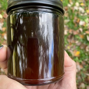 An Amber colored glass 8 oz jar candle is held in Teresas Hand  The jar is shown from the reverse without a label.  The candle is topped with a matte black lid.