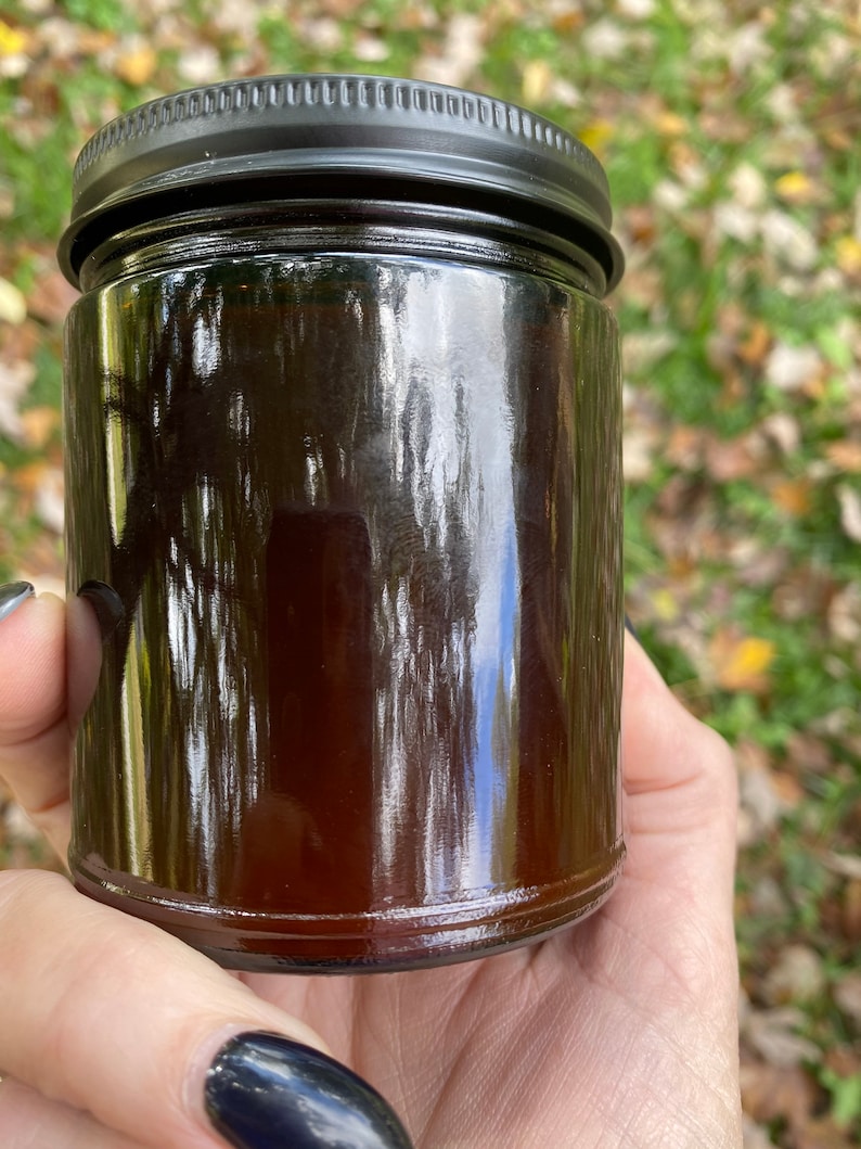 An Amber colored glass 8 oz jar candle is held in Teresas Hand  The jar is shown from the reverse without a label.  The candle is topped with a matte black lid.