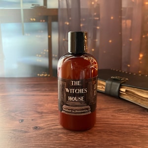 An Amber colored bottle of body wash labeled Witches House, crafted in Monrovia,I n sits on a wooden surface with a grimoire next to it. Behind the bottle is a sheer covered window with twinkly lights.