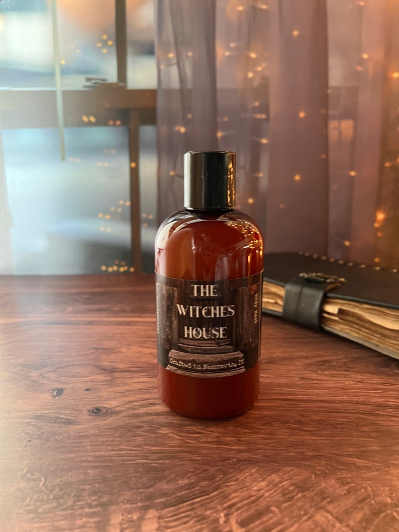 An Amber colored bottle of body wash labeled Witches House, crafted in Monrovia,I n sits on a wooden surface with a grimoire next to it. Behind the bottle is a sheer covered window with twinkly lights.