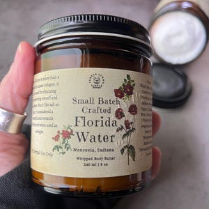 Pode incluir: Um frasco de vidro castanho com tampa preta contendo manteiga corporal batida. O r&oacute;tulo cor de creme apresenta o texto "Small Batch Crafted Florida Water", "Monrovia, Indiana" e "240 ml | 8 oz", juntamente com ilustra&ccedil;&otilde;es de flores vermelhas.