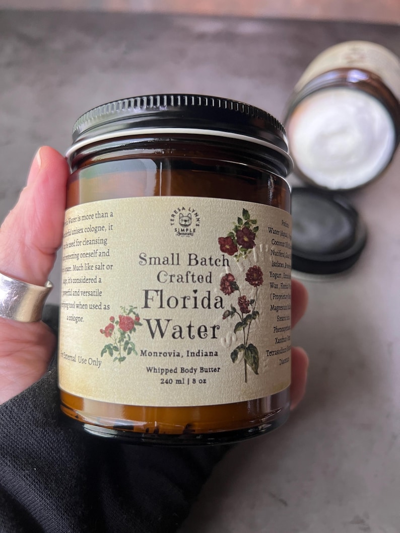 Pode incluir: Um frasco de vidro castanho com tampa preta contendo manteiga corporal batida. O r&oacute;tulo cor de creme apresenta o texto "Small Batch Crafted Florida Water", "Monrovia, Indiana" e "240 ml | 8 oz", juntamente com ilustra&ccedil;&otilde;es de flores vermelhas.