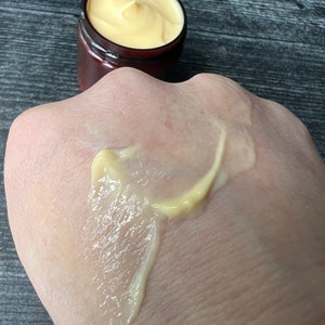 Op de afbeelding: Een klein potje met een bruin deksel bevat gele cr&egrave;me, naast een hand met een streep van dezelfde cr&egrave;me. De cr&egrave;me heeft een gladde textuur, waarschijnlijk een huidverzorgingsproduct. Het potje is klein, wat duidt op een sample of reisformaat.