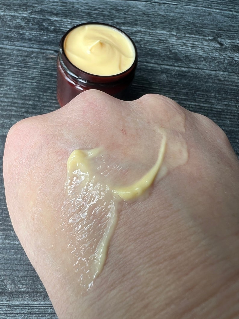 Op de afbeelding: Een klein potje met een bruin deksel bevat gele cr&egrave;me, naast een hand met een streep van dezelfde cr&egrave;me. De cr&egrave;me heeft een gladde textuur, waarschijnlijk een huidverzorgingsproduct. Het potje is klein, wat duidt op een sample of reisformaat.