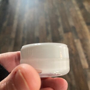 Op de afbeelding: Een kleine, ronde, witte cosmetische pot van kunststof met een schroefdeksel. De houder heeft een gladde, ondoorzichtige witte afwerking op het deksel en een licht doorschijnende witte basis, ontworpen voor het bewaren van cr&egrave;mes of lotions.
