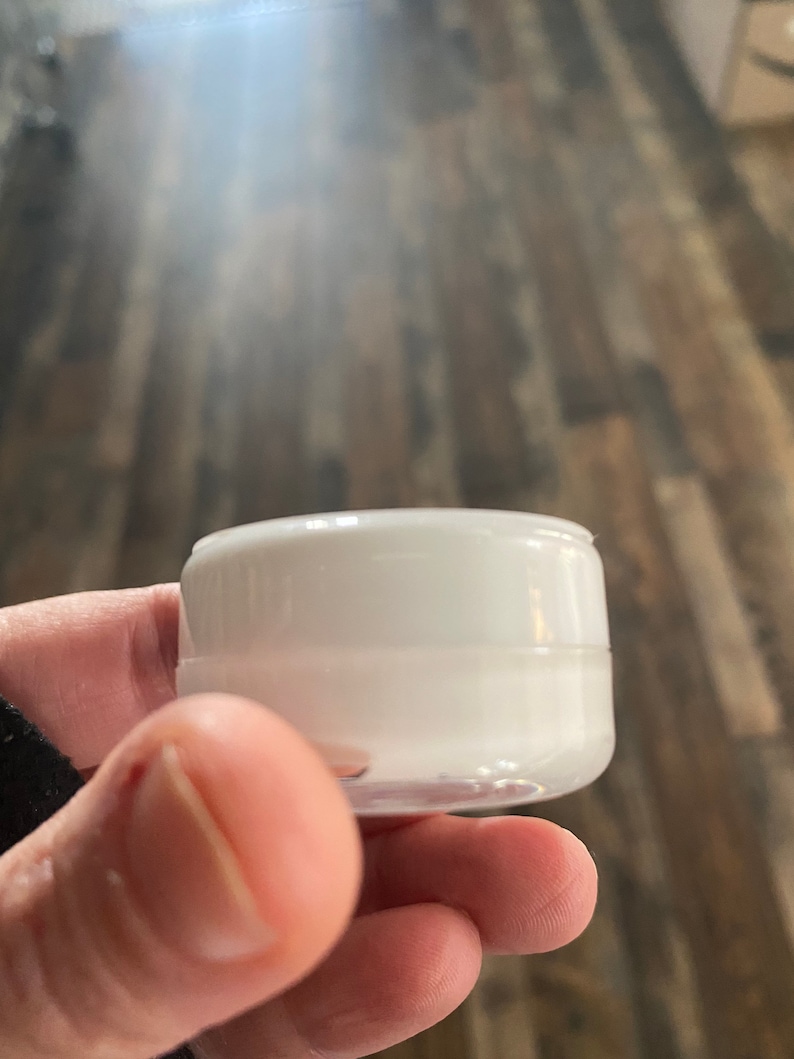 Op de afbeelding: Een kleine, ronde, witte cosmetische pot van kunststof met een schroefdeksel. De houder heeft een gladde, ondoorzichtige witte afwerking op het deksel en een licht doorschijnende witte basis, ontworpen voor het bewaren van cr&egrave;mes of lotions.