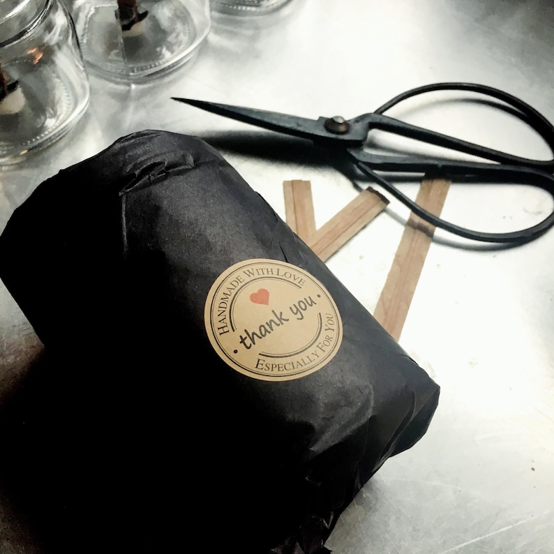 Wrapped jar A candle jar wrapped in black tissue paper with a circular Kraft   paper sticker.  Next to the wrapped candle are wooden candle wicks and vintage scissors on a graphite table.