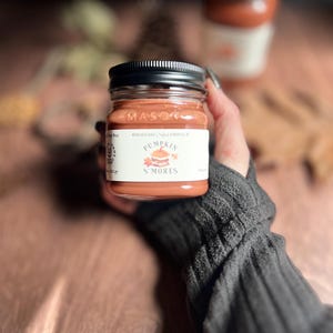 May include: A small, clear glass jar with a black lid, filled with orange-colored wax. The label reads "Pumpkin S'mores." The jar is held in a person's hand, with a second jar in the background. The jar is 3.5 inches tall.
