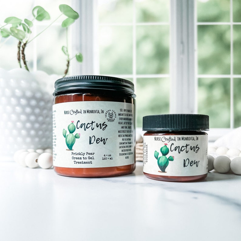 Op de afbeelding: Twee glazen potten met Cactus Dew Prickly Pear Cream to Gel Treatment. De grotere pot is 120 ml. Elke pot heeft een zwart deksel en een etiket met een cactusillustratie en de tekst "Nurse Crafted in Monrovia, IN."