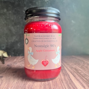 May include: A red candle in a glass jar with a black lid. The label reads "Nostalgic 90's Apple Cinnamon" and features illustrations of geese. The candle is 12 oz.