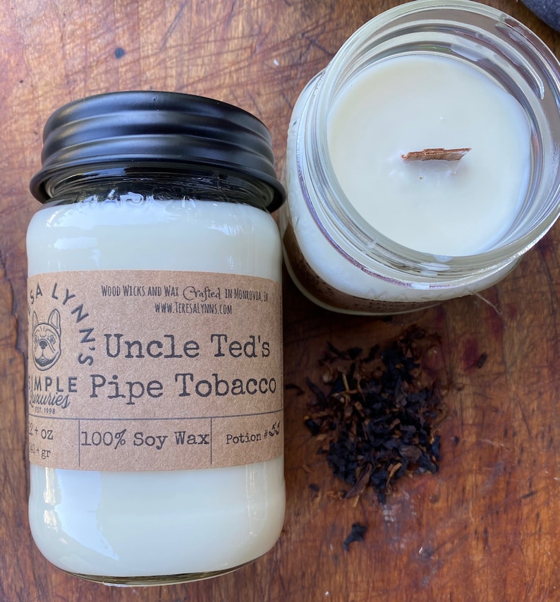 A white mason jar candle with a black lid sits on a wooden board with tobacco spilled out.  Candle is labeled "UncleTed's Pipe Tobacco" a wooden wick