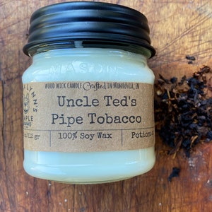 May include: A white candle in a glass jar with a black lid. The label reads "Uncle Ted's Pipe Tobacco" and "100% Soy Wax".