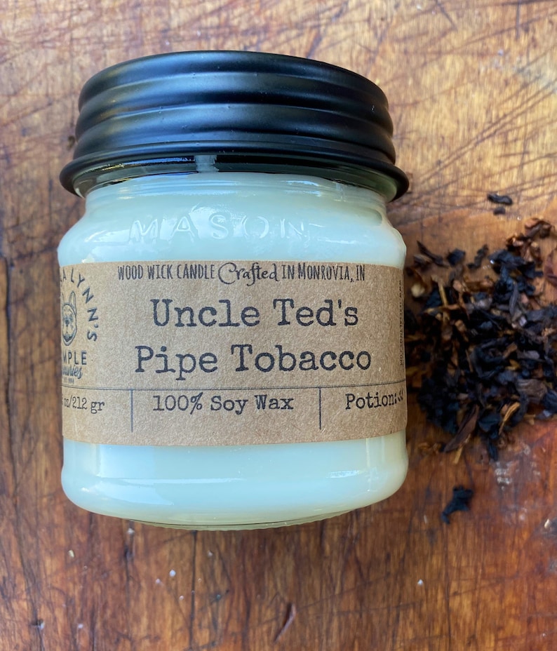 May include: A white candle in a glass jar with a black lid. The label reads "Uncle Ted's Pipe Tobacco" and "100% Soy Wax".