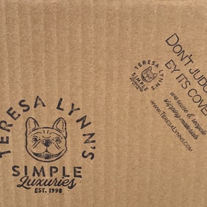 May include: A close-up of a corrugated cardboard box with printed text and logos. The box features the phrase "DON'T JUDGE A BOX BY ITS COVER." and a logo with a French bulldog. The brand name "TERESA LYNN'S SIMPLE LUXURIES" is also visible.