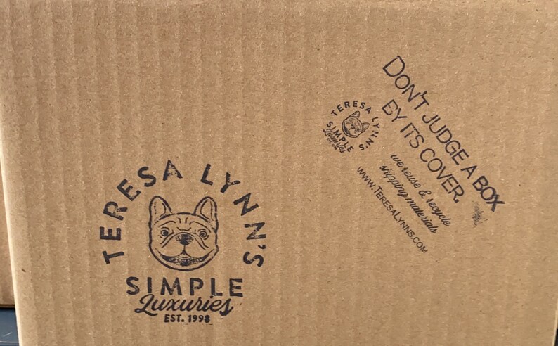 May include: A close-up of a corrugated cardboard box with printed text and logos. The box features the phrase "DON'T JUDGE A BOX BY ITS COVER." and a logo with a French bulldog. The brand name "TERESA LYNN'S SIMPLE LUXURIES" is also visible.