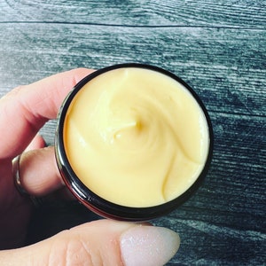 Op de afbeelding: Een klein potje gevuld met gele cr&egrave;me met een gladde, gewervelde textuur. Het potje heeft een zwarte buitenkant en een zilveren rand. De achtergrond is een grijs houten oppervlak. Het potje wordt door een persoon vastgehouden.
