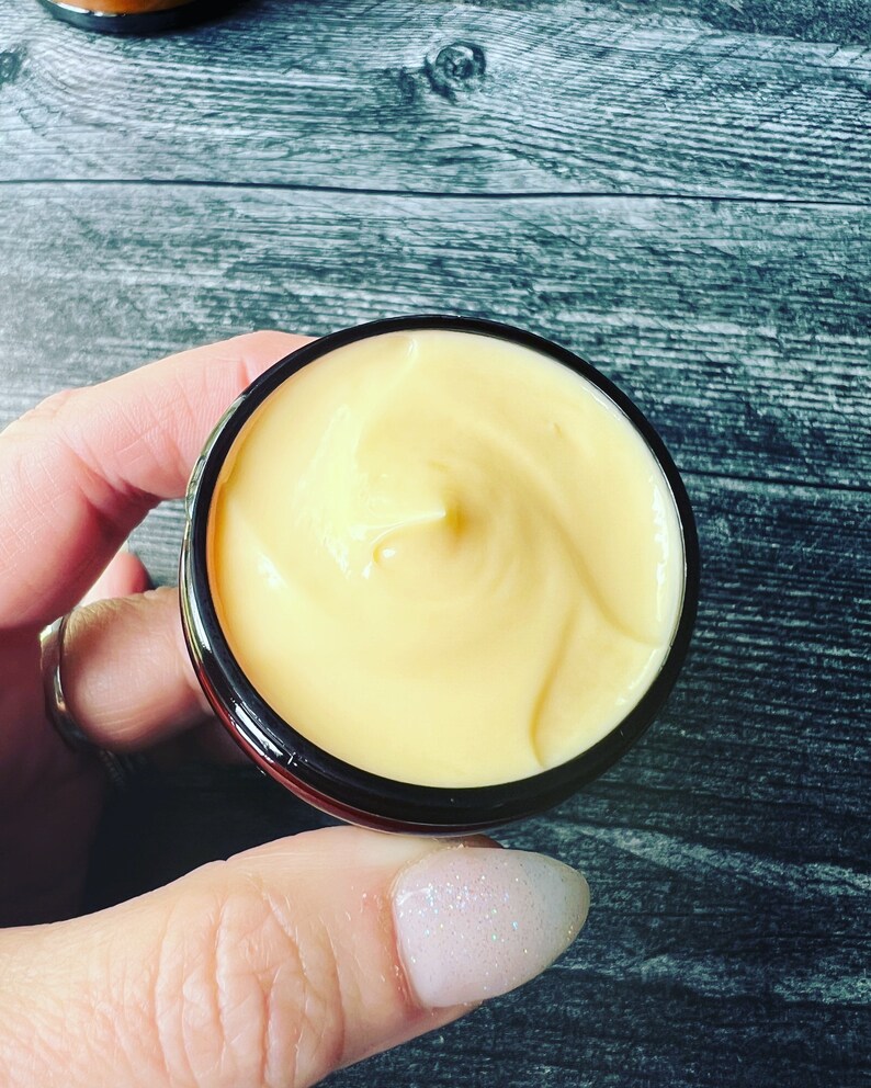 Op de afbeelding: Een klein potje gevuld met gele cr&egrave;me met een gladde, gewervelde textuur. Het potje heeft een zwarte buitenkant en een zilveren rand. De achtergrond is een grijs houten oppervlak. Het potje wordt door een persoon vastgehouden.