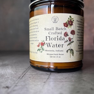 Pode incluir: Um frasco de vidro &acirc;mbar com tampa preta contendo manteiga corporal batida. O r&oacute;tulo diz Small Batch Crafted Florida Water, Monrovia, Indiana, e 240 ml, com ilustra&ccedil;&otilde;es bot&acirc;nicas de flores vermelhas e folhas verdes num fundo creme.