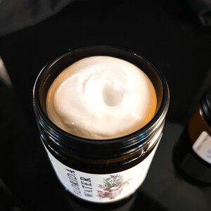 Pode incluir: Um frasco de vidro &acirc;mbar escuro preenchido com um creme branco espesso. O r&oacute;tulo no frasco apresenta uma ilustra&ccedil;&atilde;o bot&acirc;nica e o texto "FLORIDA WATER" em letras pretas. O creme tem uma textura suave e espiralada vis&iacute;vel na abertura.