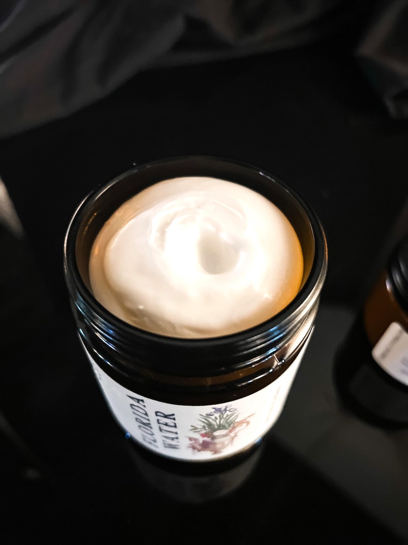 Pode incluir: Um frasco de vidro &acirc;mbar escuro preenchido com um creme branco espesso. O r&oacute;tulo no frasco apresenta uma ilustra&ccedil;&atilde;o bot&acirc;nica e o texto "FLORIDA WATER" em letras pretas. O creme tem uma textura suave e espiralada vis&iacute;vel na abertura.