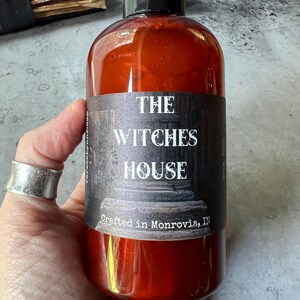 Puede incluir: Una botella de pl&aacute;stico transparente de color &aacute;mbar con tapa negra, que contiene gel de ba&ntilde;o l&iacute;quido. La etiqueta presenta un fondo oscuro y texturizado con el texto blanco "THE WITCHES HOUSE" y "Crafted in Monrovia, IN" en una fuente estilizada.
