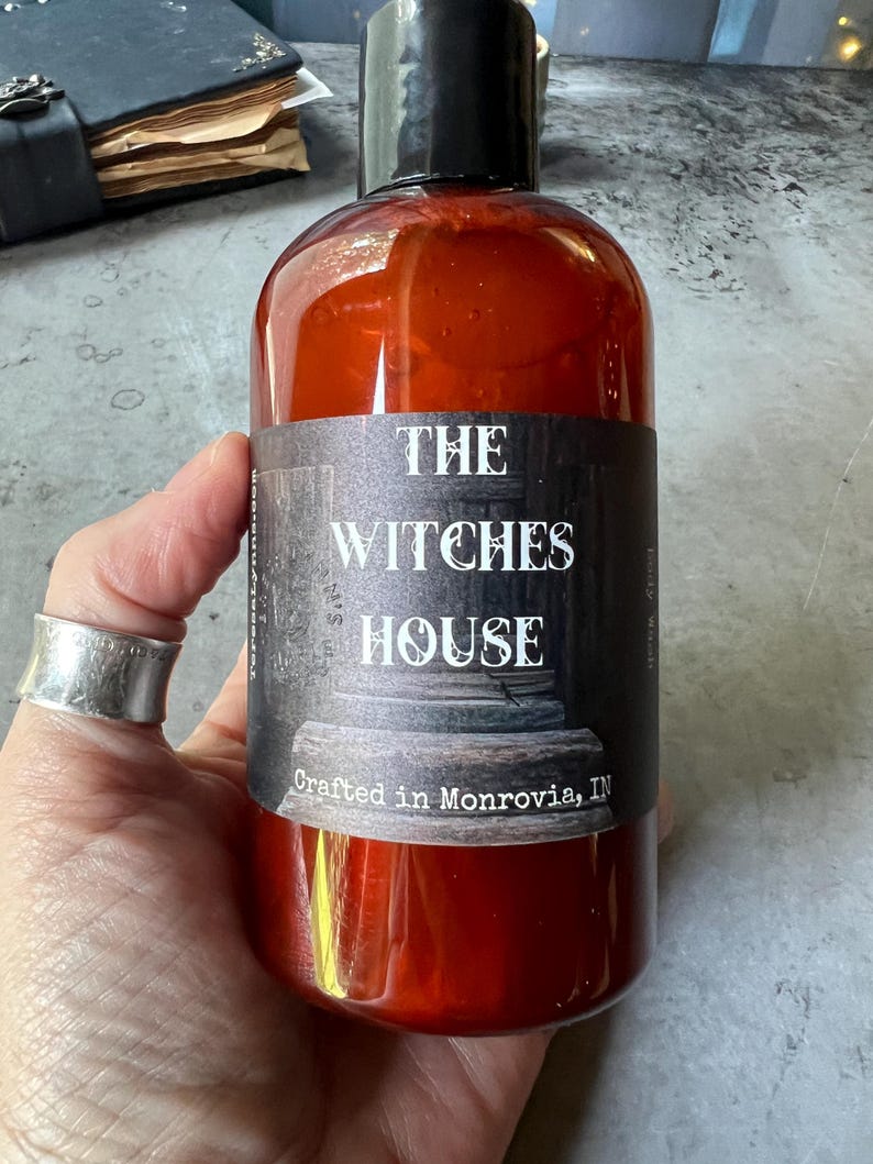 Puede incluir: Una botella de pl&aacute;stico transparente de color &aacute;mbar con tapa negra, que contiene gel de ba&ntilde;o l&iacute;quido. La etiqueta presenta un fondo oscuro y texturizado con el texto blanco "THE WITCHES HOUSE" y "Crafted in Monrovia, IN" en una fuente estilizada.
