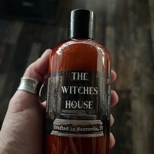 An Amber colored bottle of body wash labeled Witches House, crafted in Monrovia, In is held in Teresas hand.
