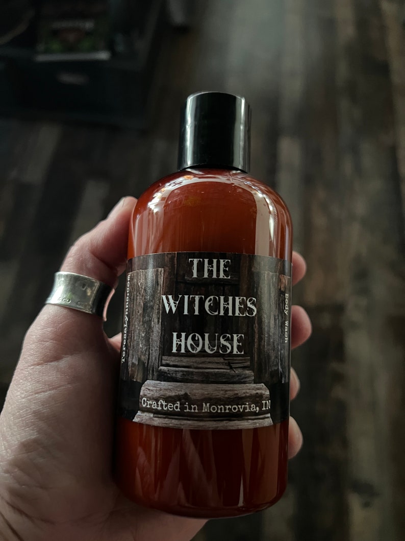 An Amber colored bottle of body wash labeled Witches House, crafted in Monrovia, In is held in Teresas hand.