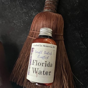 Op de afbeelding: Een amberkleurige glazen fles Florida Water met een zilveren schroefdop en een wit etiket met de tekst Crafted in Monrovia, IN, Small Batch Crafted, Florida Water, geplaatst voor een bruine strooien handveger met een met draad omwikkelde steel.