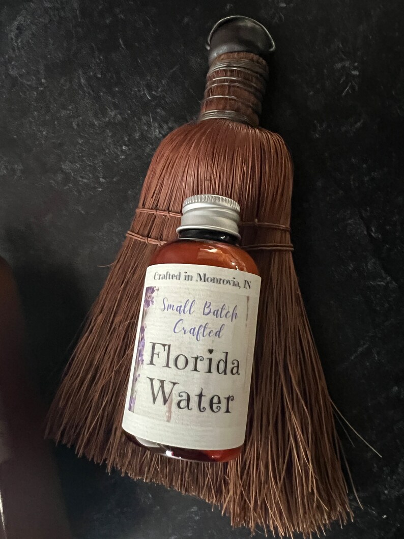 Op de afbeelding: Een amberkleurige glazen fles Florida Water met een zilveren schroefdop en een wit etiket met de tekst Crafted in Monrovia, IN, Small Batch Crafted, Florida Water, geplaatst voor een bruine strooien handveger met een met draad omwikkelde steel.