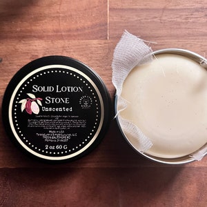 以下が含まれることがあります： 「Solid Lotion Stone Unscented」の文字とカカオポッドのグラフィックが入った黒い丸い缶。その隣には、布で包まれた固形ローションストーンが入った銀色の缶があります。ローションストーンの重量は60gです。