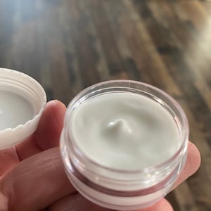 Op de afbeelding: Een kleine, ronde, doorzichtige plastic pot gevuld met een gladde, witte, ondoorzichtige cr&egrave;me. De witte schroefdop is losgemaakt en aan de linkerkant geplaatst, waardoor de dikke, romige textuur van het product in de pot zichtbaar is.