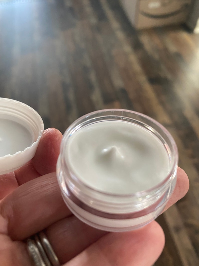 Op de afbeelding: Een kleine, ronde, doorzichtige plastic pot gevuld met een gladde, witte, ondoorzichtige cr&egrave;me. De witte schroefdop is losgemaakt en aan de linkerkant geplaatst, waardoor de dikke, romige textuur van het product in de pot zichtbaar is.