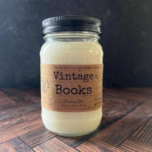 May include: A glass jar candle with a black lid and a brown label. The label reads "Vintage Books" and lists ingredients. The candle contains 12 oz of wax. The jar sits on a wooden surface.