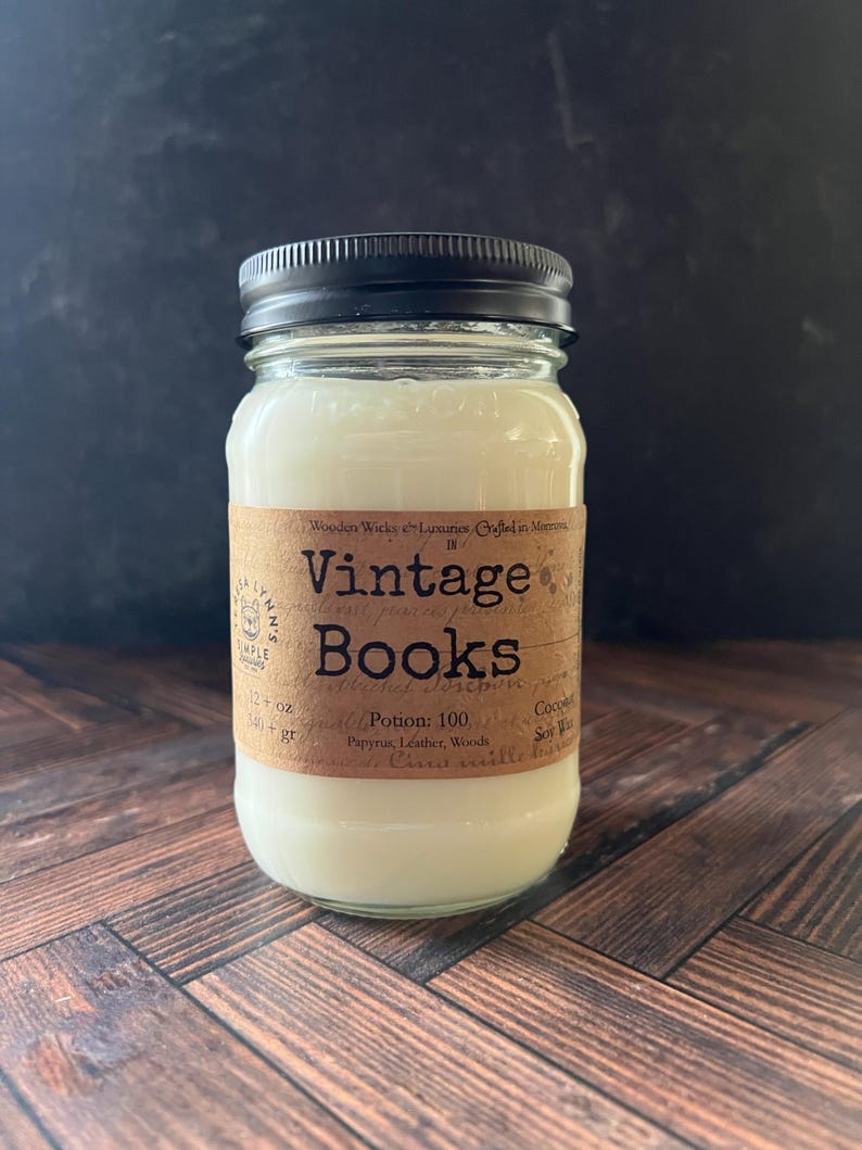 May include: A glass jar candle with a black lid and a brown label. The label reads "Vintage Books" and lists ingredients. The candle contains 12 oz of wax. The jar sits on a wooden surface.