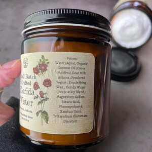 Pode incluir: Um frasco de vidro &acirc;mbar com tampa preta contendo manteiga corporal batida. O r&oacute;tulo apresenta uma ilustra&ccedil;&atilde;o bot&acirc;nica de flores vermelhas e uma lista de ingredientes incluindo &aacute;gua, &oacute;leo de coco org&acirc;nico, leite de cabra e mistura Florida Water.