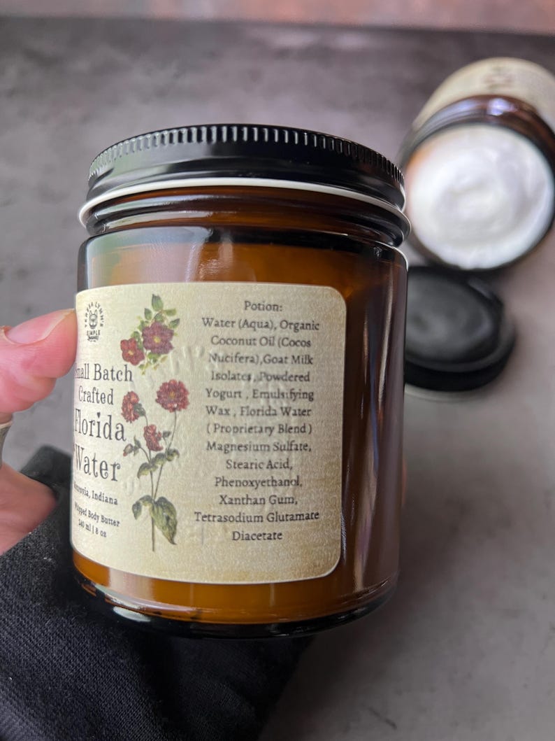 Pode incluir: Um frasco de vidro &acirc;mbar com tampa preta contendo manteiga corporal batida. O r&oacute;tulo apresenta uma ilustra&ccedil;&atilde;o bot&acirc;nica de flores vermelhas e uma lista de ingredientes incluindo &aacute;gua, &oacute;leo de coco org&acirc;nico, leite de cabra e mistura Florida Water.
