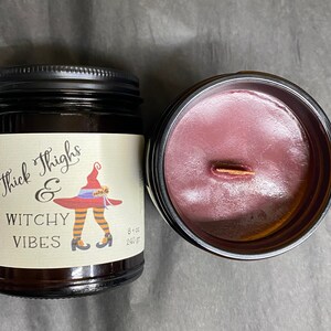 A dark candle jar.  The label reads Thick Thighs & Witchy Vibes.  The graphic is anime inspired witch legs with black and orange striped stockings, topped with red witch hat.  An open candle shows dark cherry red wax and a wooden wick.