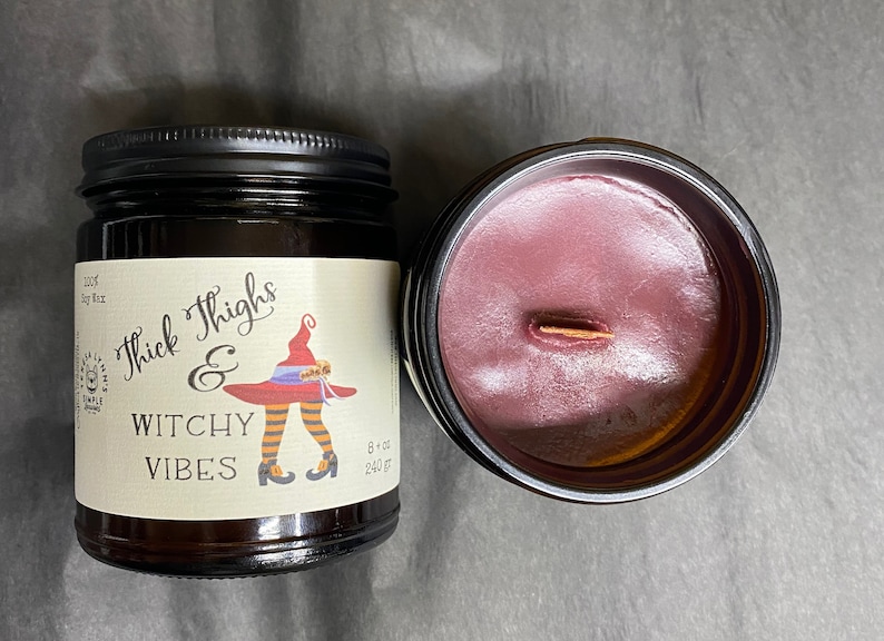 A dark candle jar.  The label reads Thick Thighs & Witchy Vibes.  The graphic is anime inspired witch legs with black and orange striped stockings, topped with red witch hat.  An open candle shows dark cherry red wax and a wooden wick.
