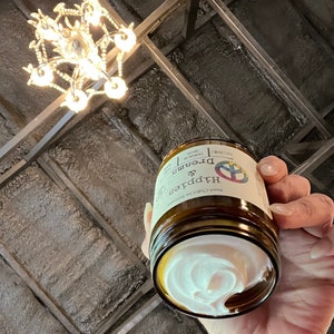 May include: A brown glass jar of Dreams & Hippies hand cream, held in a hand. The jar has a white label with text and a peace sign logo. The cream inside is white and swirled. A chandelier hangs in the background.