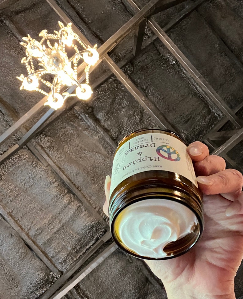 May include: A brown glass jar of Dreams & Hippies hand cream, held in a hand. The jar has a white label with text and a peace sign logo. The cream inside is white and swirled. A chandelier hangs in the background.