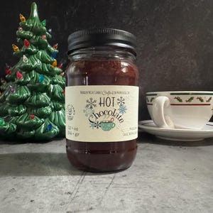 May include: A glass jar candle with a black lid and a cream-colored label featuring the text HOT Chocolate, snowflake graphics, and a small mug illustration. The label indicates a volume of 12 oz and 354 gr, with text stating Wooden Wick Candle.