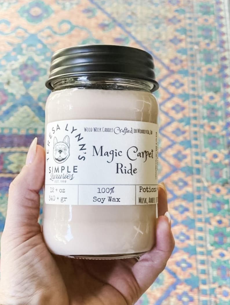 A hand holds a jar candle labeled Magic Carpet Ride with a Vintage Rug in the background. Label reads 100% Soy Wax Musk, Amber, Oud