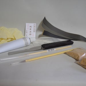 Puede incluir: Un kit para reparar artículos de cuero, que contiene un tubo de adhesivo, una espátula pequeña, dos palitos de madera, una pequeña bolsa de polvo marrón, un par de guantes blancos, una pequeña cinta de cinta negra y una pequeña tarjeta con texto japonés.