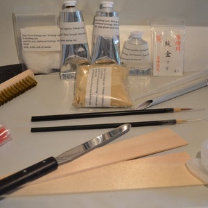 May include: A collection of kintsugi repair supplies, including tubes of silver-colored paste, brushes, sandpaper, and small packets of powder. Also included are tools such as a spatula, brushes, and wooden boards. The kit is designed for traditional kintsugi repair.